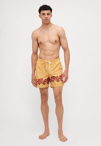 Badeshorts aus gelbem Stoff mit einem wellenförmigen roten Muster am Saum, ausgestattet mit einem elastischen Bund mit Kordelzug und einer entspannten Passform.