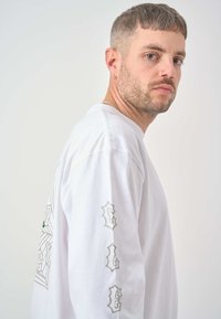 Weißes Langarm-T-Shirt mit grafischem Design und grünen Akzenten auf der Brust. Verfügt über graue "G"-Buchstaben auf dem unteren Arm. Weicher Stoff.
