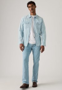 Ανοιχτό μπλε τζιν πουκάμισο με δύο τσέπες στο στήθος, φορεμένο πάνω από λευκό μπλουζάκι, συνδυασμένο με ανοιχτό μπλε τζιν παντελόνια και σκούρα καφέ μπότες, σε όρθια στάση.