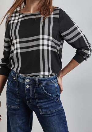 Vrouw draagt een zwart-grijs geruit shirt met lange mouwen, ingestopt in een donkerblauwe spijkerbroek, met één hand op haar heup.