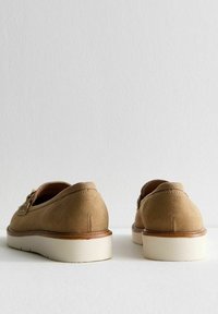 Chaussures en suede marron avec une forme arrondie, dotées d'un talon bas et d'une semelle en caoutchouc couleur crème. Comprend des éléments décoratifs sur le côté.