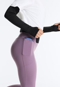 Oseba z rokavi iz črne tkanine in belim zgornjim delom, ki nosi vijoličaste legice s stranskimi žepi, drži modri pametni telefon.