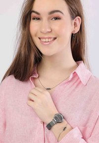 Ronde zwarte horloge met mesh band, gedragen om een pols. Model in een lichtroze gestreept shirt, met een zilveren ketting en oorbellen.