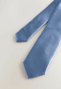 Cravate en soie texturée bleu clair posée à plat sur une surface blanche, montrant les extrémités larges et étroites.