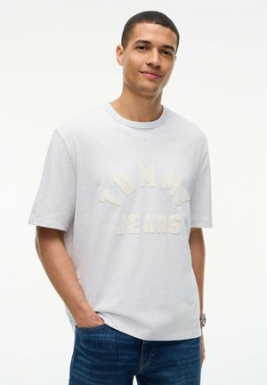 Junger Mann mit kurzen Haaren, der ein weißes Tommy Jeans T-Shirt und blaue Jeans trägt, steht mit einer Hand in der Tasche vor weißem Hintergrund.