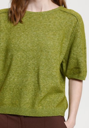 Femme portant un pull en tricot vert à manches courtes avec un ourlet côtelé et un pantalon marron, une main dans la poche.