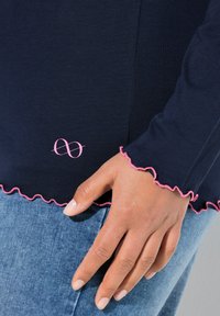 Námořnická modrá dlouhá halenka s růžovým volánkem na spodním lemu a odpovídajícími manžetami. Obsahuje růžové nekonečné logo poblíž lemu. Materiál vypadá měkký a přiléhavý.