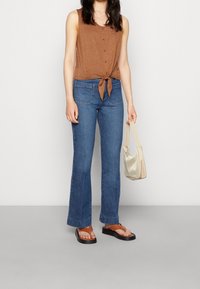 Bruine mouwloze top met knopen en strikdetail, gecombineerd met flared blauwe denim jeans, bruine sandalen en een lichtgekleurde schoudertas.