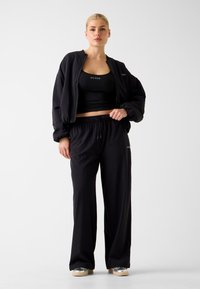 Tenue de sport noire composée d'une veste bomber courte et d'un pantalon à cordon de serrage. Tissu lisse, coupe décontractée, avec des accents de logo sur le haut et le pantalon.