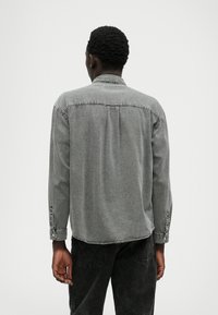 Camisa de denim cinza com mangas longas, punhos com botões, textura suave e um ajuste relaxado. Inclui um detalhe na parte de trás e costuras discretas.