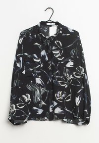 Camicia nera a maniche lunghe con un motivo floreale bianco e grigio, due tasche frontali sul petto, appesa a una gruccia nera.