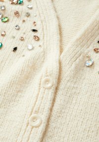 Cardigan couleur crème avec un tricot texturé, deux gros boutons, et des embellissements scintillants dispersés de différentes formes et couleurs.