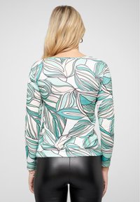 Haut à manches longues blanc avec un motif de feuilles teal et noir. Il présente un col rond et une coupe ajustée. Mélange de coton, texture lisse.