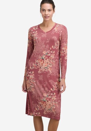 Kvinde iført en støvet lyserød knælængde kjole med lange ærmer og blomsterprint, der står mod en ensfarvet hvid baggrund.