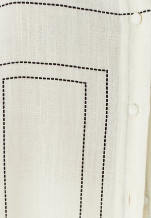 Bela tkanina z črnim prešitekom, ki tvori pravokotnike, z gumbom za zapiranje. Mehka tekstura, lahka in rahlo prosojna.