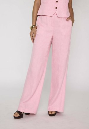 Donna che indossa pantaloni rosa chiaro a gamba larga e gilet senza maniche abbinato, accessoriata con bracciale dorato e tacchi neri aperti.