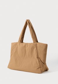 Suuri pehmeä vaaleanruskea kangaskassi leveillä pehmustetuilla kahvoilla, joka seisoo pystyssä valkoista taustaa vasten.