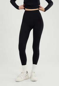 Osoba ubrana w czarne legginsy z wysokim stanem, białe skarpetki i białe trampki, stojąca na jasnym tle.