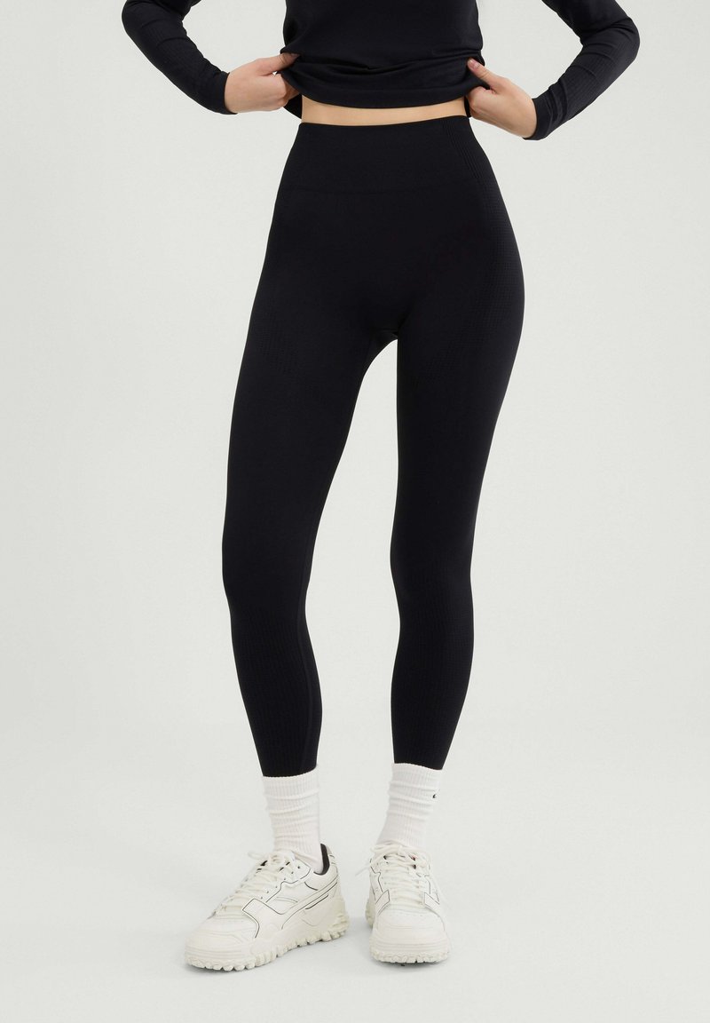 Osoba ubrana w czarne legginsy z wysokim stanem, białe skarpetki i białe trampki, stojąca na jasnym tle.