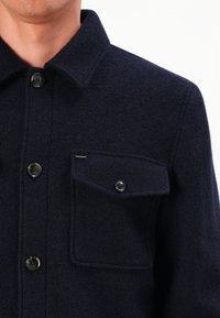 Mann in einem dunkelblauen Wollmantel mit Kragen, drei sichtbaren Knöpfen und einer geknöpften Brusttasche vor einem weißen Hintergrund.