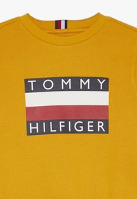 Sudadera amarilla de algodón, con un gráfico rectangular que presenta "TOMMY" encima de "HILFIGER" en blanco y negro, con una franja roja y blanca debajo.
