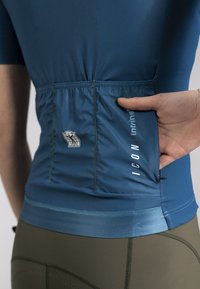 Blue fitted cycling jersey with a gathered back pocket, featuring logo detail and contrasting bottom hem. Made from lightweight, stretch fabric.