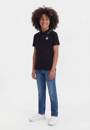 Smilende barn med krøllet hår iført en sort polo shirt, blå jeans og hvide sneakers, stående mod en ensfarvet hvid baggrund.