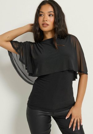 Vrouw met lang donker haar, gekleed in een doorschijnende zwarte cape top en zwarte leren broek, poserend met één hand achter haar hoofd.