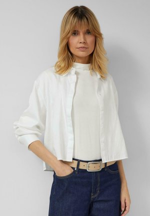 Biała koszula na guziki z podwiniętymi rękawami, noszona na jasnej bluzce. Ciemnoniebieskie dżinsy i beżowy pasek z prostokątną klamrą.