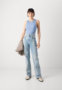 Hellblau gestreiftes Tanktop, hoch geschnittene verblasste blaue Jeans, hält einen karierten Stoff. Trägt creamfarbene Schuhe mit schwarzen Akzenten.