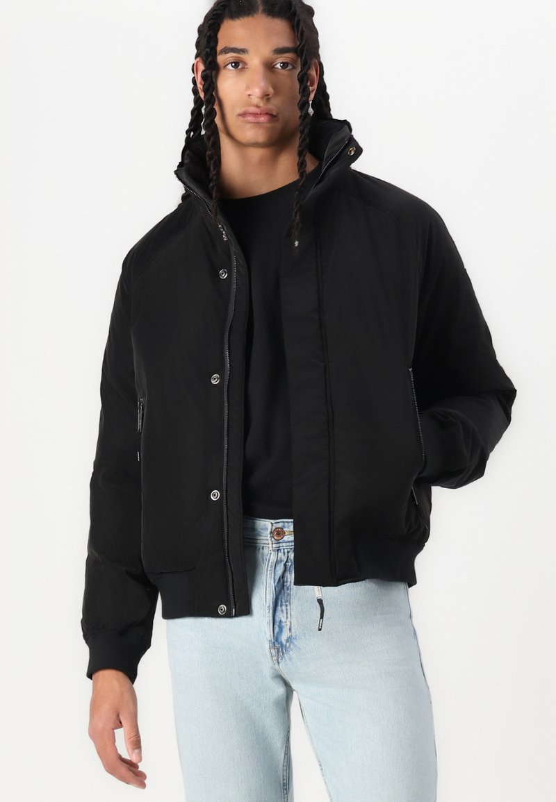 Chaqueta bomber negra con cremallera, botones a presión, puños elásticos y bolsillos laterales, hecha de tela suave, llevada sobre una camiseta negra y unos vaqueros.