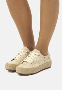 Zapatilla beige con diseño perforado, suela de estilo espadrille tejida, cierre de cordones y suela de goma. Decorada con pequeños agujeros decorativos.