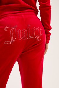 Červené velúrové tepláky s veľkým, trblietavým kamienkovým zdobením so slovom "Juicy" na zadnej strane. Hladký povrch, elastický pás.