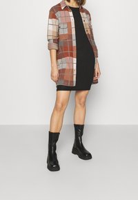 Giacca camicia a quadri marrone e grigia sopra un vestito nero, abbinata a stivali alla caviglia con suola spessa nera. Presenta maniche arrotolate e design con bottoni.