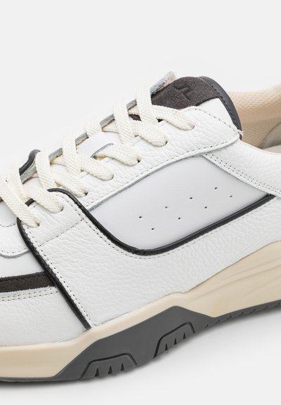 White leather sneaker with textured upper, cream laces, black accents, and a chunky beige sole. Features perforated detail for ventilation.