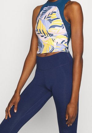 Femme portant un haut court sans manches avec des motifs de feuilles jaunes, bleues et roses, associé à un legging bleu marine taille haute.