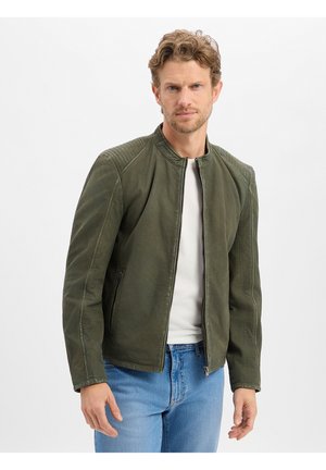 Mann mit hellbraunen lockigen Haaren, der eine geöffnete olivgrüne Jacke über einem weißen Hemd und blauen Jeans trägt, vor einem schlichten Hintergrund.