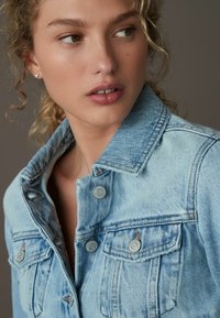 Chaqueta de mezclilla azul desgastada con botones plateados, dos bolsillos en el pecho y costuras visibles, llevada sobre una blusa de color sólido.