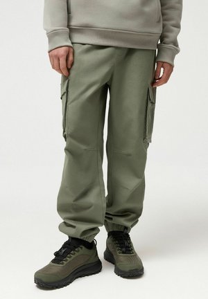 Mężczyzna ubrany w oliwkowo zielone spodnie bojówki z bocznymi kieszeniami, czarne trampki i szarą bluzę, stojący na białym tle.