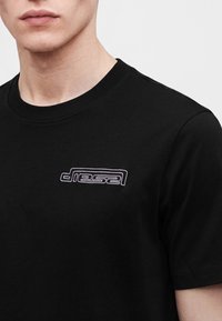 Κοντινό πλάνο ενός ατόμου που φοράει μαύρο t-shirt με λευκό περίγραμμα λογότυπου "Diesel" στο αριστερό μέρος του στήθους και με στρογγυλή λαιμόκοψη.