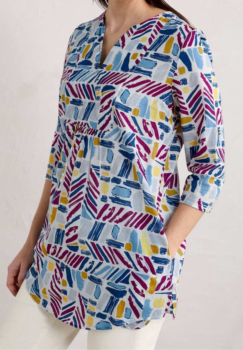 Una blusa azul claro con escote en V, que presenta patrones abstractos multicolores en rosa, amarillo y azul. Tiene mangas largas y bolsillos laterales.