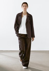 Maroon trench coat over a white top; high-waisted, wide-leg trousers in brown. Model wears white shoes, standing against a neutral background.