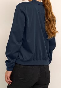 Veste bomber bleu marine à manches longues, poignets et ourlet côtelés ; texture lisse ; dotée d'un col rond et de détails de couture minimalistes.