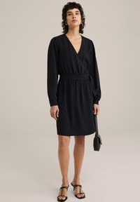 Černé zavinovací šaty s dlouhými rukávy, texturovanou tkaninou, elastickým pasem a vázaným detailem. V páru s malou černou crossbody taškou.