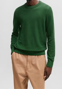 Pull en maille verte avec un col rond et des poignets côtelés, associé à un pantalon beige clair. Le tissu a un motif texturé.