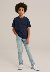 Jūras spēku īsā piedurkne T-krekls ar krūšu kabatu, savienots ar gaiši ziliem džinsiem un baltām sporta apaviem. Modelis stāv ar rokām kabatās, neitrālā fonā.