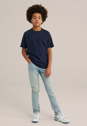 Camiseta de manga corta azul marino con un bolsillo en el pecho, combinada con jeans de un azul claro y zapatillas blancas. El modelo está de pie con las manos en los bolsillos, sobre un fondo neutro.