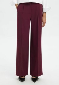 Femme portant un pantalon bordeaux taille haute à jambes larges avec des plis avant, associé à des talons noirs à bout pointu et un haut blanc à manches longues.