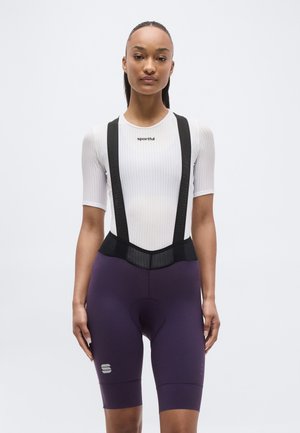 Γυναίκα που φοράει λευκή ποδηλατική μπλούζα με κοντά μανίκια και μαύρο βερμούδα με τιράντες, στέκεται μπροστά σε απλό φόντο.