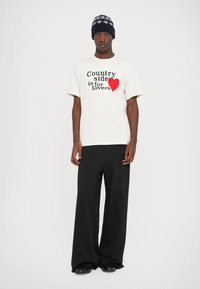 Man in een wit T-shirt met de tekst "Het platteland is voor geliefden" en een rode hart, zwarte wijde broek en een blauwe gebreide hoed.